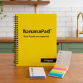 A yellow BananaPad Original hands-on productivity organiser notebook stands on a wooden countertop in a kitchen setting, with two tasks strips.